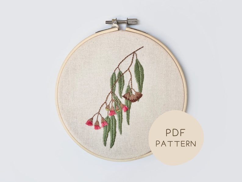 May include: Embroidered eucalyptus branch with green leaves and pink flowers, displayed in a wooden embroidery hoop. The design is stitched on a beige fabric. A circular label with the text "PDF PATTERN" is visible.