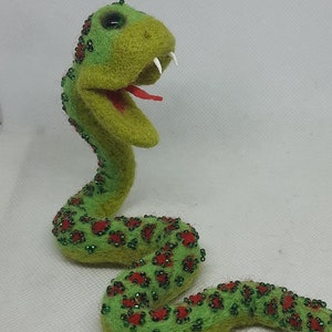 May include: A green felt snake with red and green bead accents. The snake is curled up with its head raised and mouth open.