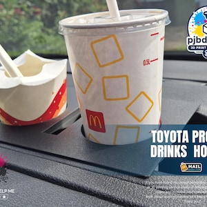 May include: A black plastic Toyota Proace drinks holder featuring two circular cutouts. One cutout holds a white paper cup with yellow square patterns and a red logo, while the other holds a white paper container with a white plastic spoon inside.