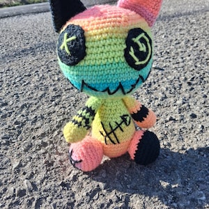 May include: A handmade crochet doll in a cat-like design. The doll features a rainbow color scheme, black accents, and stitched details. The eyes are black with yellow cross stitches, and the mouth is a stitched grin.