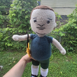 May include: A handmade crochet doll with a light skin tone, brown hair, and a dark blue shirt. The doll is wearing green shorts, white socks, and black and white shoes. It is holding a yellow object with a black strap.