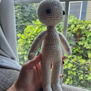 May include: A beige crochet doll with a rounded head, slender arms, and legs. The doll features two small, round, black plastic buttons for eyes, positioned on the upper portion of the head. The entire body is crafted from yarn in a consistent stitch.