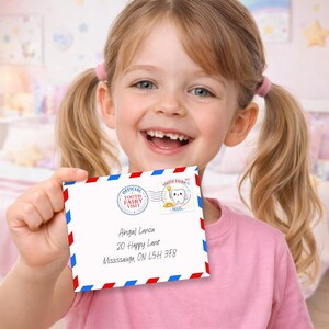 Puede incluir: Una niña sonriente sostiene un sobre blanco con bordes rojos y azules. El sobre tiene el texto "Official Tooth Fairy Visit" y una dirección. La niña lleva una camiseta rosa y tiene coletas rubias.