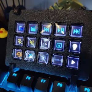 May include: A black keyboard with a small, black, rectangular device attached to it. The device has 12 buttons with various icons, including a play button, a pause button, a fast forward button, a rewind button, a mute button, a volume button, a brightness button, and a 1% button. The buttons are illuminated with blue and white lights.