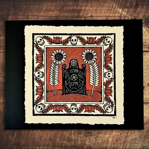 May include: A black and red linocut print featuring a central tombstone design with the dates 1728 and 1791. The print is framed by a border of skulls, flowers, and stylized leaves. The artwork is on textured paper.