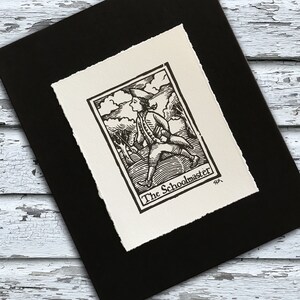 May include: A black and white woodblock print of a man in a hat and coat walking through a forest. The print is titled "The Schoolmaster" and is framed in a black frame.