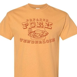 May include: Mustard-colored t-shirt with a brown graphic. The graphic features the words "BREADED PORK TENDERLOIN" above and below an illustration of a sandwich, a can, and the outline of a state.