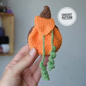 May include: A small orange crochet pumpkin pouch with a brown stem and two green curly tendrils hanging down. The pouch features a flap closure and is labeled with a circular white graphic containing the text CROCHET PATTERN in black.