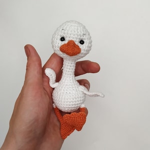 May include: A white crocheted goose toy with orange feet and black eyes. The goose is sitting in a hand.