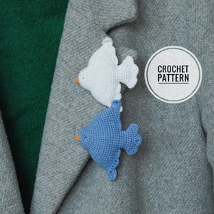 Peut inclure: Deux broches en forme d'oiseaux au crochet, une blanche et une bleue, épinglées sur un manteau gris. Les oiseaux ont des becs oranges. Un cercle blanc avec les mots "CROCHET PATTERN" est dans le coin supérieur droit.