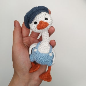 May include: A crocheted goose toy wearing a blue hat and blue overalls. The goose has white feathers, orange feet, and black eyes.