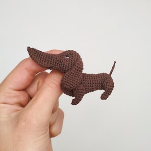 May include: A brown crocheted dachshund toy with black eyes. The toy is being held by a hand.