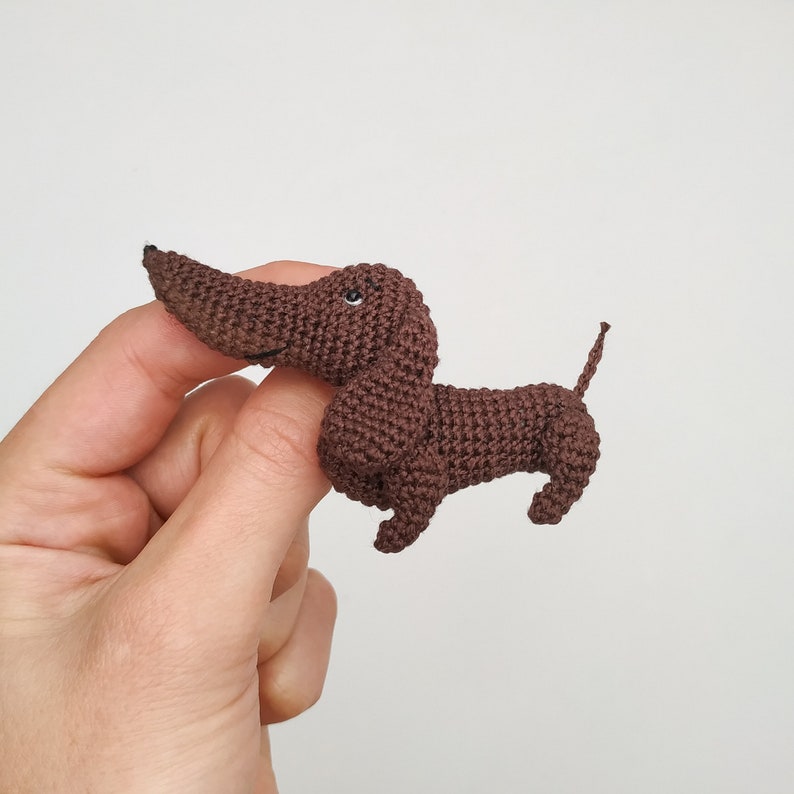 May include: A brown crocheted dachshund toy with black eyes. The toy is being held by a hand.