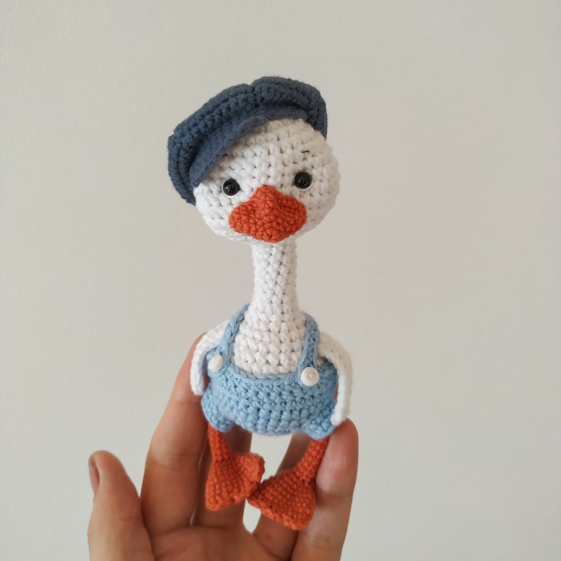 May include: A white crocheted goose wearing a blue hat and blue overalls. The goose has orange feet and black eyes.