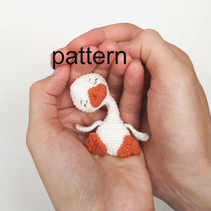 Puede incluir: Un ganso blanco de crochet con patas naranjas y un ojo cerrado, sostenido en las palmas de dos manos. La palabra "pattern" está escrita sobre el ganso.