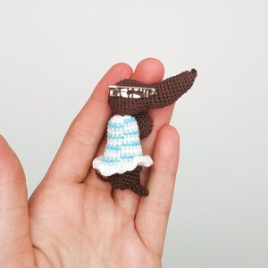 May include: A brown crocheted dachshund wearing a blue and white striped dress with a white crocheted ruffle. The dachshund is attached to a silver safety pin.
