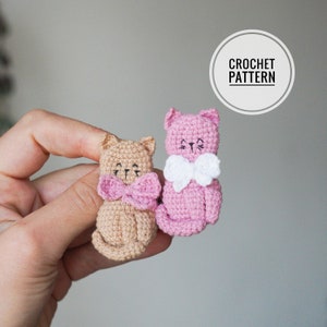 Pode incluir: Dois brinquedos de crochê em forma de gato, um rosa e outro bege, com laços brancos e rosa. Os brinquedos são pequenos e têm olhos e narizes bordados. O texto CROCHET PATTERN é visível no fundo.