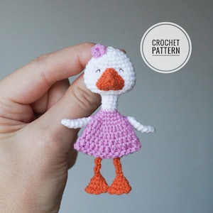 May include: A white crocheted goose wearing a pink dress and orange feet. The goose has black eyes and a small pink flower on its head. The image includes the text "CROCHET PATTERN".