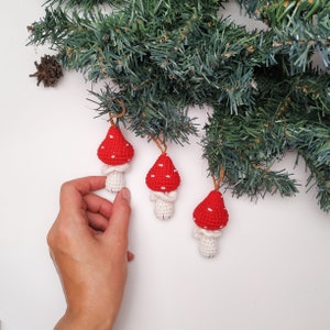 Pu&ograve; includere: Tre ornamenti a forma di fungo in crochet rosso e bianco con pois bianchi. Gli ornamenti sono appesi a un ramo di pino verde.