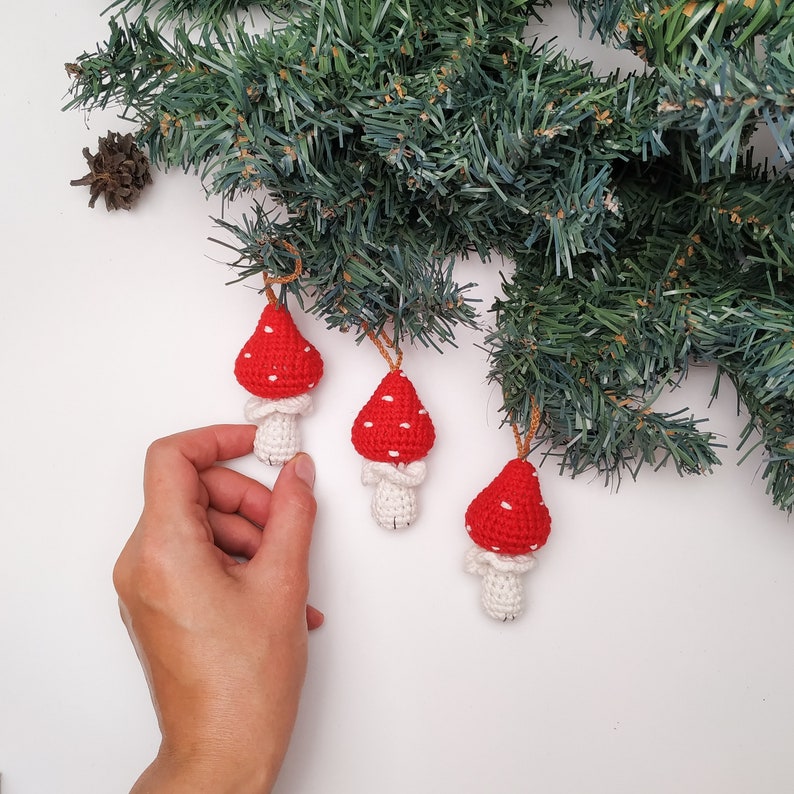 Pu&ograve; includere: Tre ornamenti a forma di fungo in crochet rosso e bianco con pois bianchi. Gli ornamenti sono appesi a un ramo di pino verde.