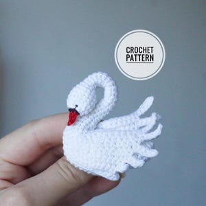 Peut inclure: Un petit cygne crocheté blanc avec un bec rouge et un détail d'œil noir. Le cygne présente un cou courbé et des plumes d'ailes texturées. Un graphique circulaire blanc dans le coin supérieur droit contient le texte CROCHET PATTERN.