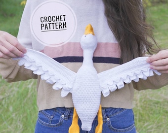 May include: A white crochet goose with yellow feet and beak, and outstretched wings. The wings have a scalloped edge. The text "CROCHET PATTERN" is in a white circle. The goose is held by a person wearing a sweater and jeans.