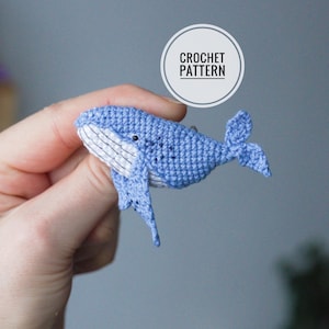 Könnte beinhalten: Eine gehäkelte blaue Walfigur mit weißen Akzenten an Bauch und Mund. Der Wal hat eine kleine Schwanzflosse und Flossen. Das Bild enthält eine runde Grafik mit der Aufschrift "CROCHET PATTERN".