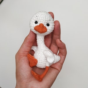 May include: A white crocheted goose with orange feet and a black beak. The goose is sitting in a hand.