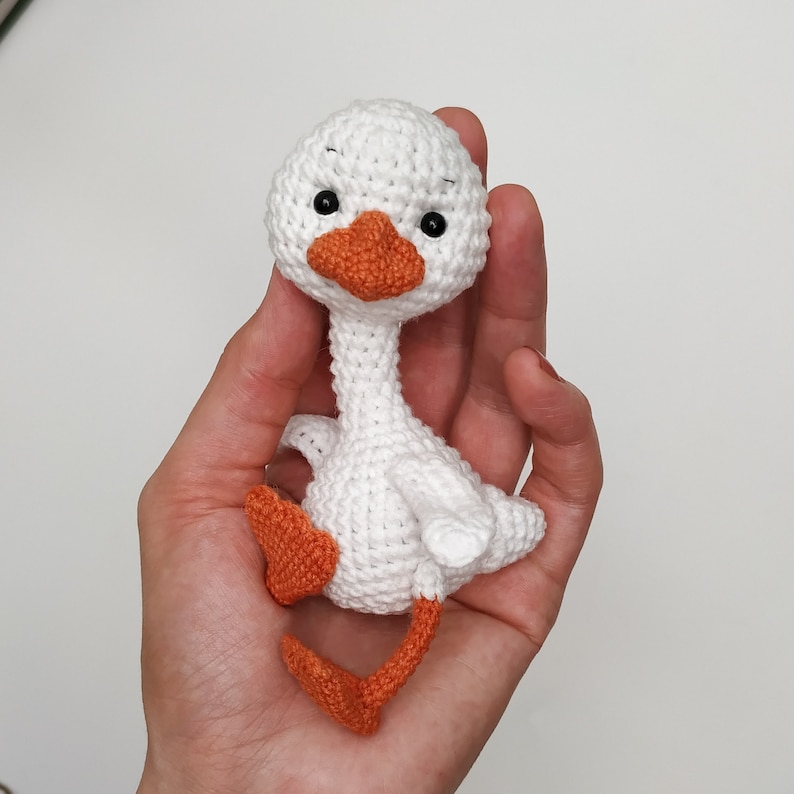 May include: A white crocheted goose with orange feet and a black beak. The goose is sitting in a hand.