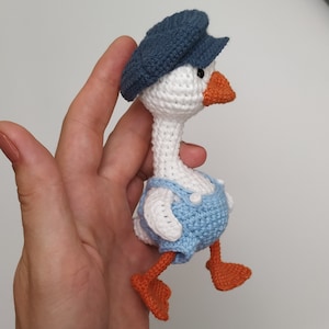 May include: A white crocheted goose wearing a blue cap and blue overalls. The goose has orange feet and a beak.