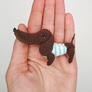 May include: A small crocheted dachshund wearing a blue and white striped shirt. The dachshund is brown and has black eyes.