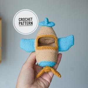 Op de afbeelding: Een gehaakt vliegtuigspeelgoed in bruin, blauw en goud. Het speelgoed heeft blauwe vleugels, een propeller en een blauwe propeller. De tekst "CROCHET PATTERN" staat in een witte cirkel.