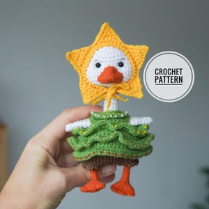 May include: A handmade crochet duck doll with a yellow star-shaped hat, white face, and orange beak and feet. The duck wears a green dress with gold accents. The doll has the text "CROCHET PATTERN" in a circle.