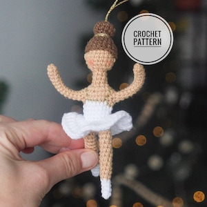 May include: Crochet pattern for a ballerina ornament. The ornament is made of white and brown yarn and is wearing a white tutu. The ornament is hanging from a gold string.