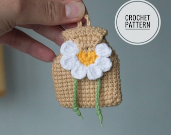 Op de afbeelding: Een kleine beige gehaakte rugzak-bedel met een wit en geel madeliefje op de voorflap en twee dunne groene garenstrengen die naar beneden hangen. De tekst CROCHET PATTERN staat in een witte cirkel in de rechterbovenhoek.