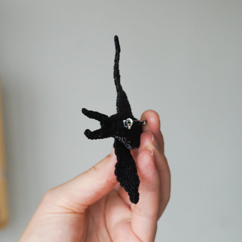 K&ouml;nnte beinhalten: Ein schwarzer, gestrickter Vogel-Ansteckpin mit silberfarbener Nadel. Der Vogel hat ausgebreitete Fl&uuml;gel und einen langen, schlanken Hals. Der Pin wird vor einem schlichten, hellgrauen Hintergrund gehalten.