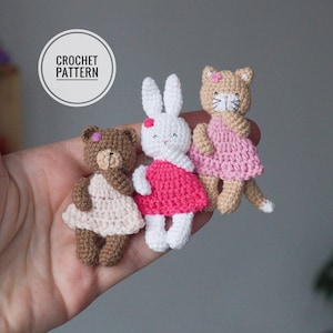 Op de afbeelding: Drie gehaakte knuffels: een bruine beer, een wit konijn en een beige kat. Elk knuffeldier draagt een roze jurk. De knuffels zitten op een hand.