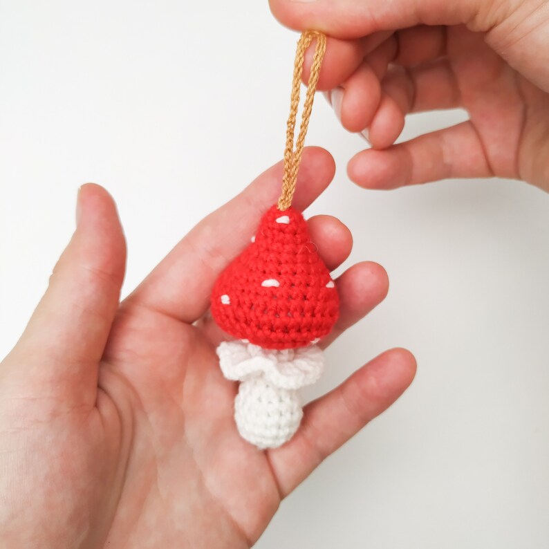 Pu&ograve; includere: Un ornamento a forma di fungo in crochet rosso e bianco con punti bianchi sul cappello rosso. Il fungo &egrave; attaccato a uno spago dorato.
