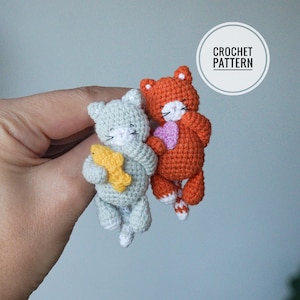 Puede incluir: Dos juguetes de crochet con forma de gato, uno gris con un pez amarillo y otro naranja con un corazón rosa. Los juguetes están siendo sostenidos por una mano. "CROCHET PATTERN" está escrito en un círculo en la esquina superior derecha de la imagen.
