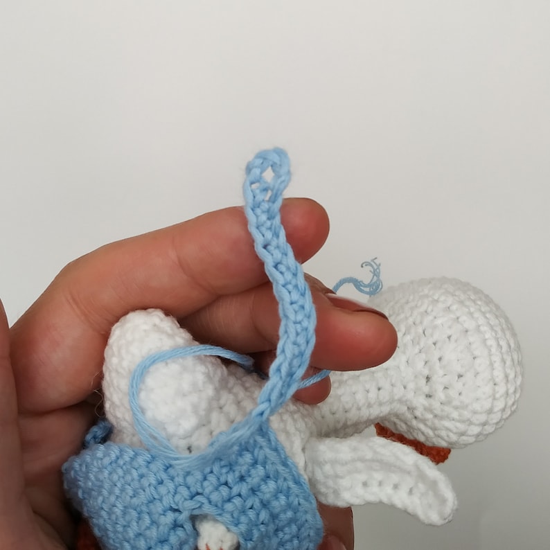 May include: A crocheted white and blue baby doll with a blue crocheted dress. The doll is being held by a hand.