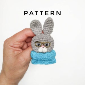May include: A handmade gray crochet bunny wearing glasses and a blue scarf. The bunny has long ears and a white muzzle. The word "PATTERN" is printed above the bunny.