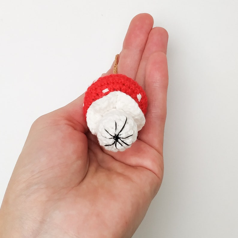 Pu&ograve; includere: Un giocattolo a forma di fungo all'uncinetto con un cappello rosso e un gambo bianco. Il fungo ha un viso ricamato nero.