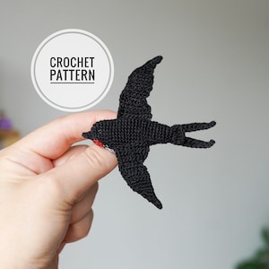 Puede incluir: Un pájaro negro de ganchillo con un detalle rojo y blanco en el pecho. El pájaro está sostenido en una mano. Un círculo blanco con las palabras "CROCHET PATTERN" está en el fondo.