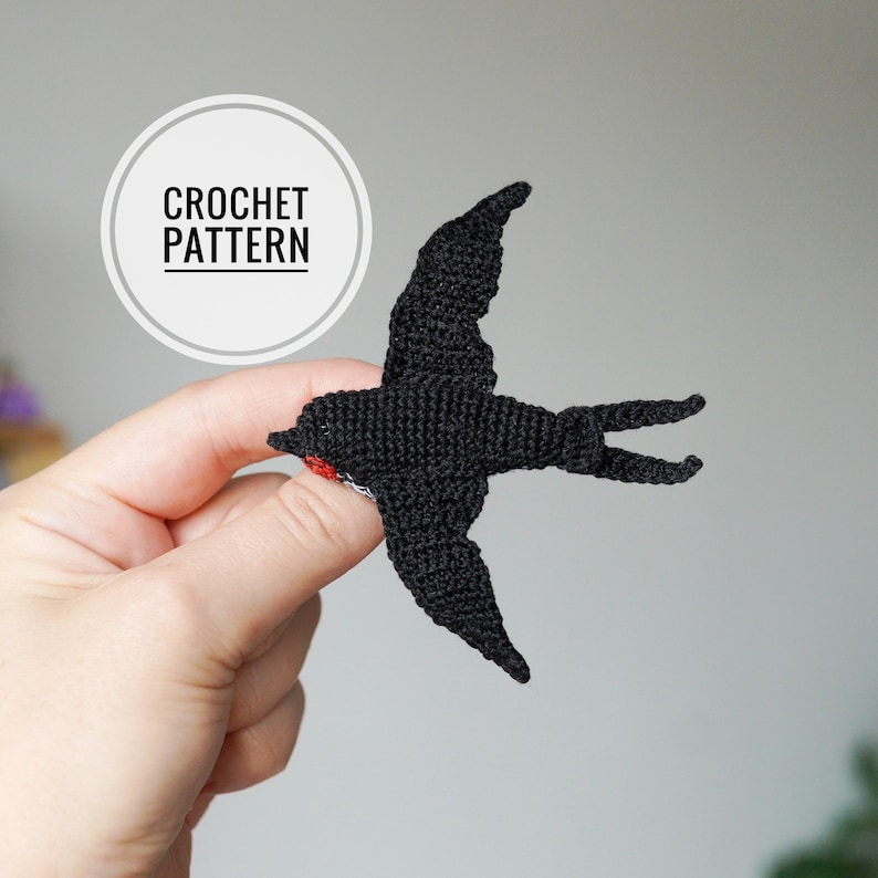 K&ouml;nnte beinhalten: Ein schwarzer H&auml;kelvogel mit rotem und wei&szlig;em Akzent auf der Brust. Der Vogel wird in einer Hand gehalten. Ein wei&szlig;er Kreis mit den Worten "CROCHET PATTERN" befindet sich im Hintergrund.