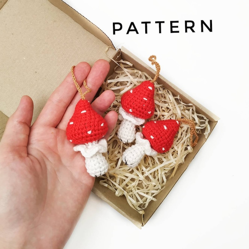 Pu&ograve; includere: Tre ornamenti a forma di fungo in crochet rosso e bianco con punti bianchi. I funghi sono in una scatola marrone con carta triturata.