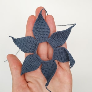 May include: A crocheted flower with six petals in a dark blue color. The flower is held in a hand, with the back of the hand facing the camera.