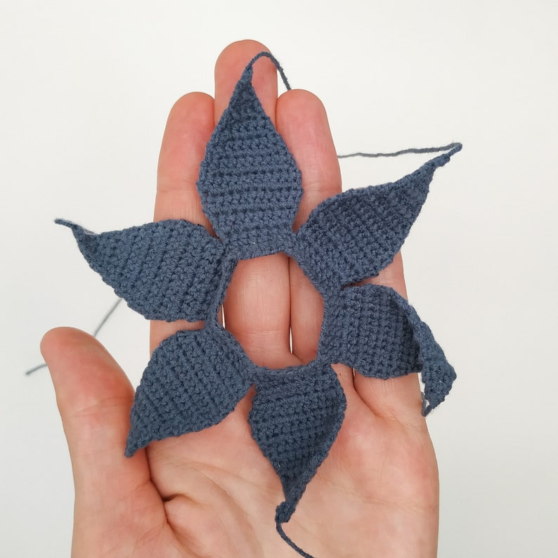 May include: A crocheted flower with six petals in a dark blue color. The flower is held in a hand, with the back of the hand facing the camera.