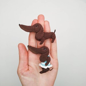 May include: Two crocheted dachshund dog figurines. One is brown and the other is brown with a white and blue dress.