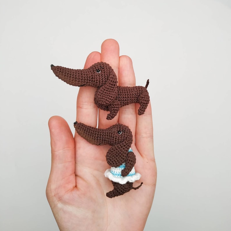May include: Two crocheted dachshund dog figurines. One is brown and the other is brown with a white and blue dress.