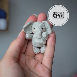 Puede incluir: Un pequeño elefante de crochet gris amigurumi con ojos negros y mejillas rosadas. El juguete está siendo sostenido en la mano de una persona. CROCHET PATTERN está escrito en texto blanco sobre un círculo blanco.